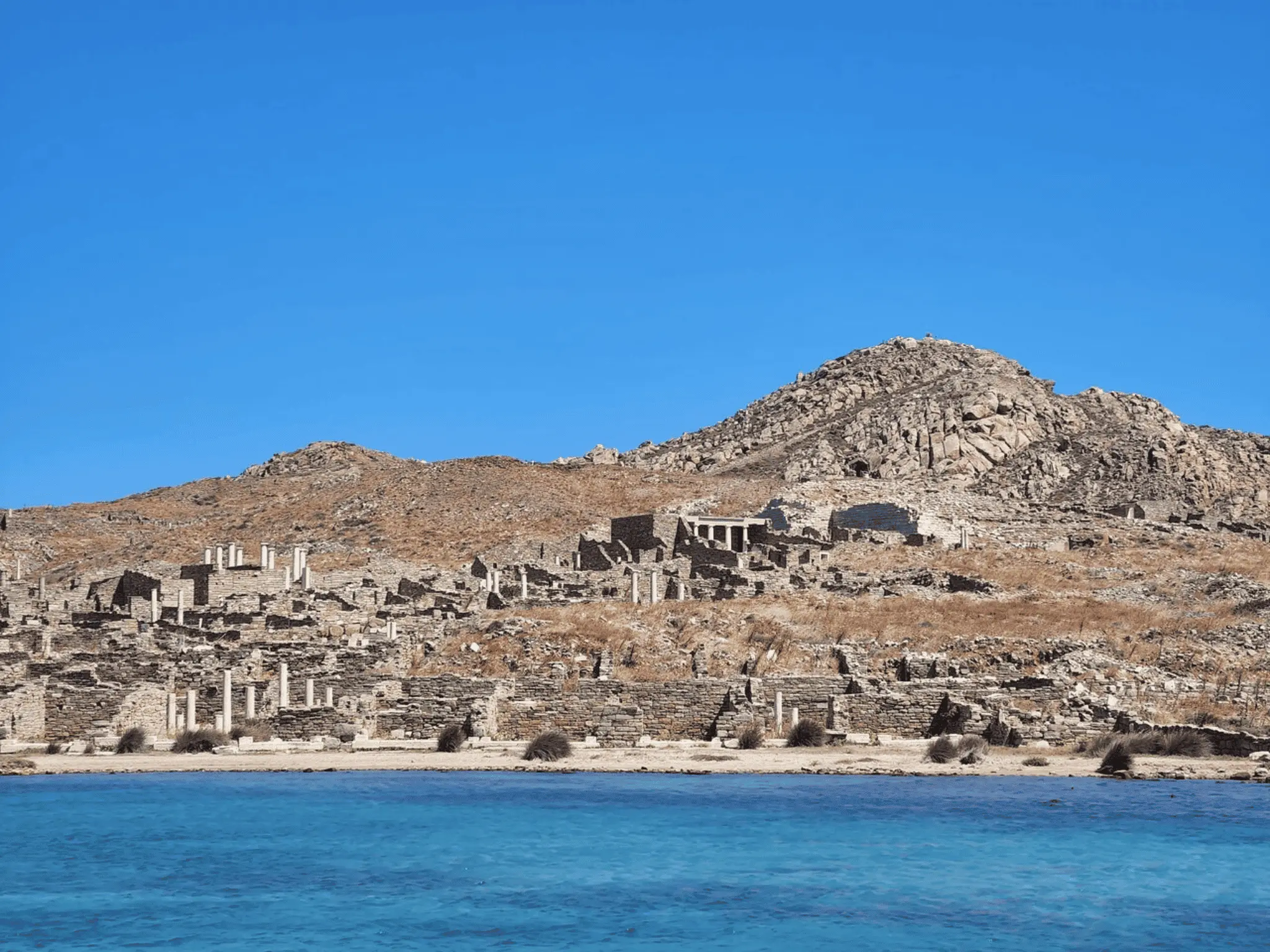 Ancient stone ruins and marble columns on a dry hillside overbooking the blue Aegean sea under a clear sky.
