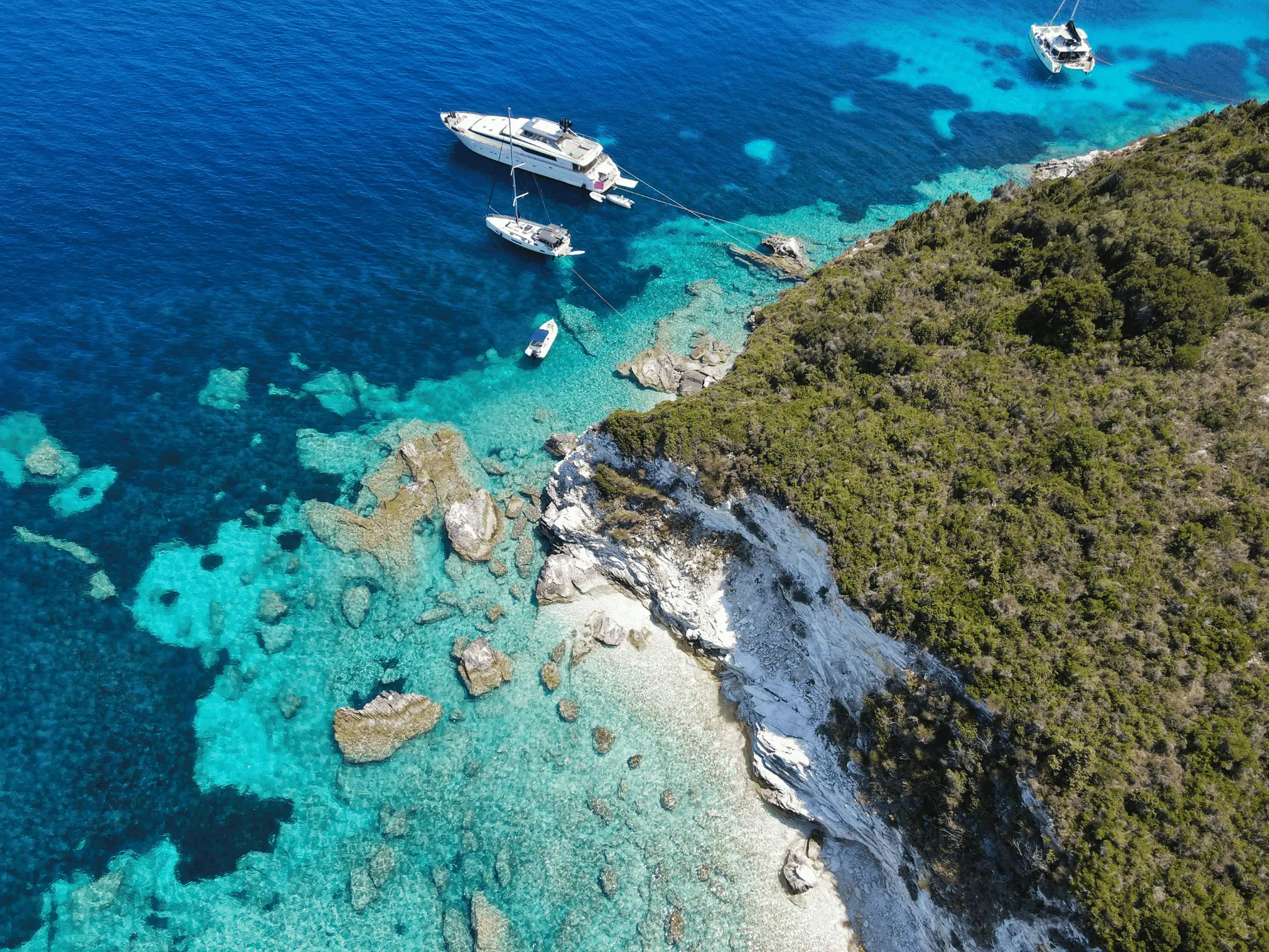 Aerial view of boats anchored in crystal-clear turquoise water along a rocky island coastline.