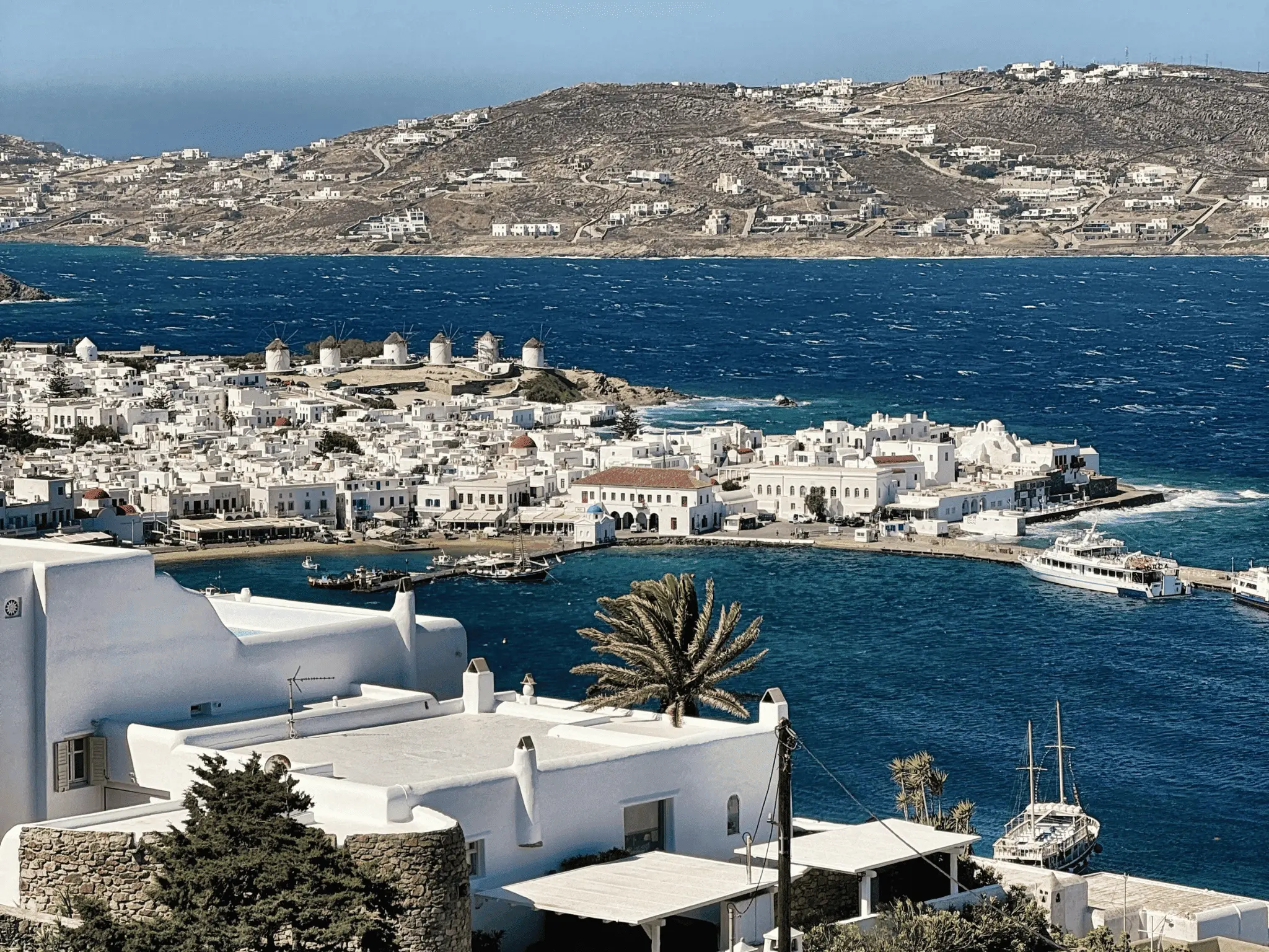 View of Mykonos harbor with whitewashed buildings, traditional windmills, boats and deep blue sea on a windy day.