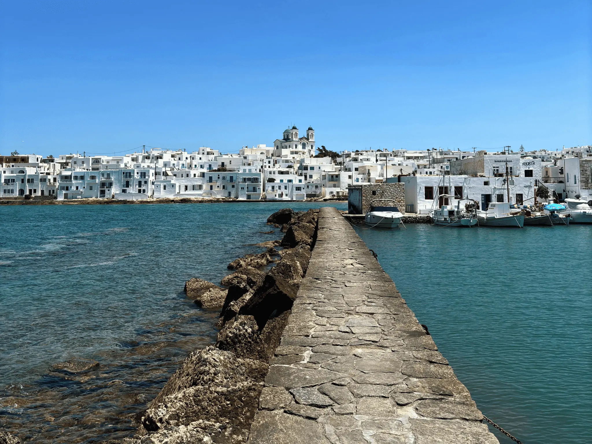Coastal view of a Greek island harbor with boats, white houses, and a stone walkway over the sea.
