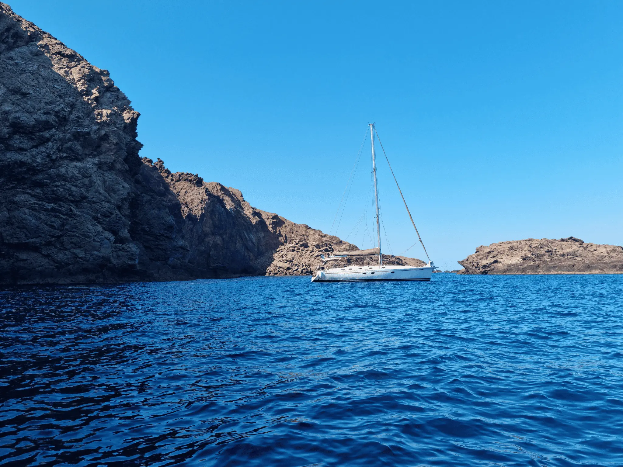 A white sailboat anchored in deep blue water between rugged rocky cliffs under a clear sky.