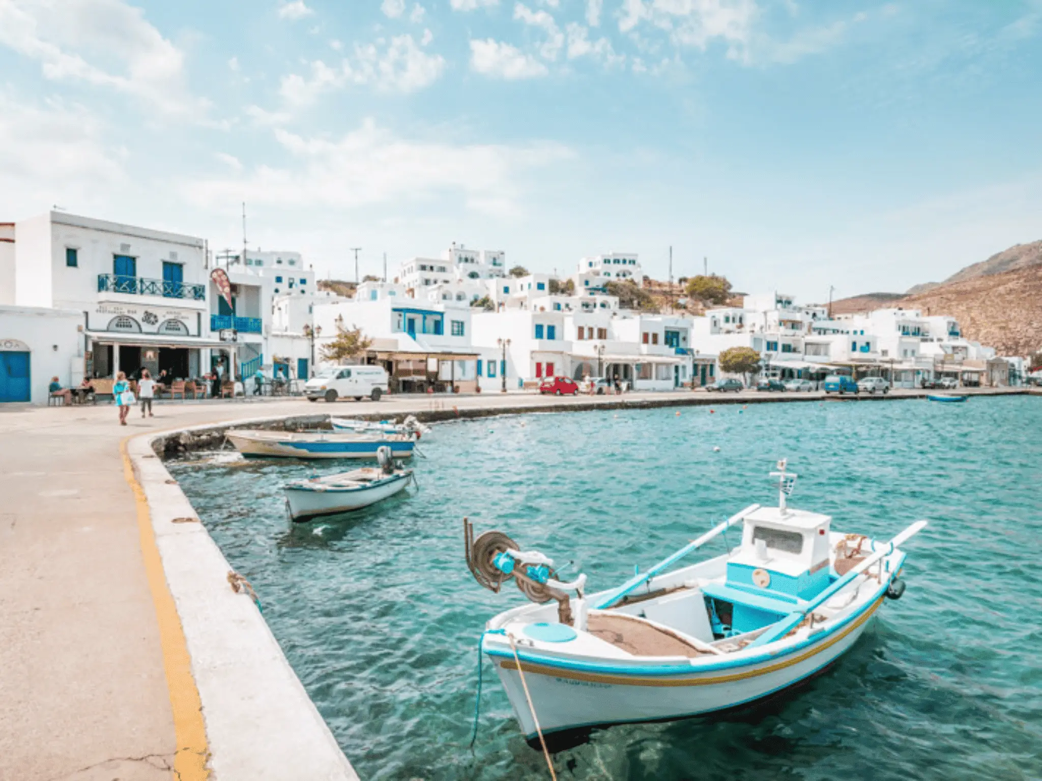 Small fishing boats moored along a seaside promenade with whitewashed buildings and blue accents in a Greek island harbor.