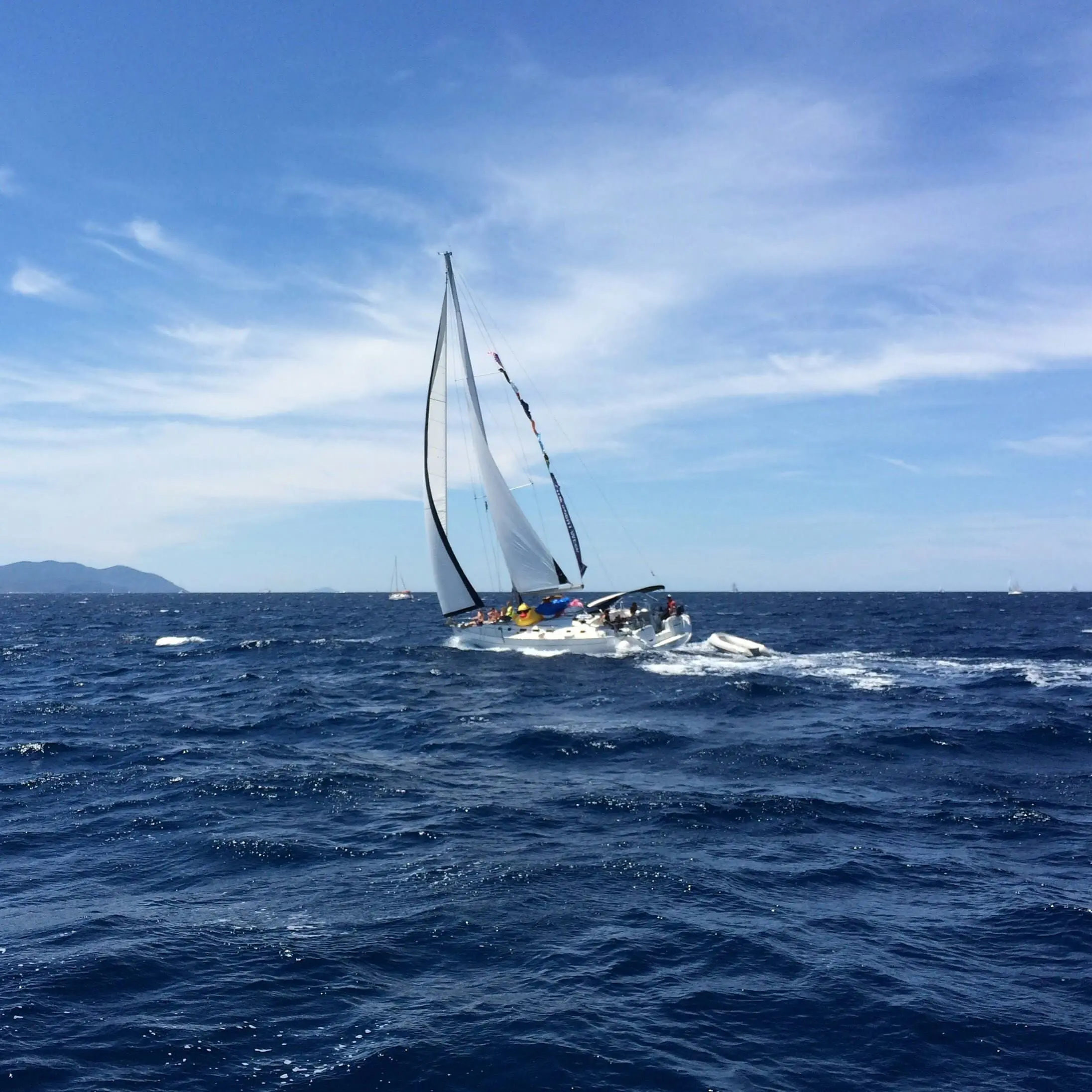 A sailing yacht with full sails glides through deep blue waters on a clear, sunny day with scattered clouds and distant mountains on the horizon.