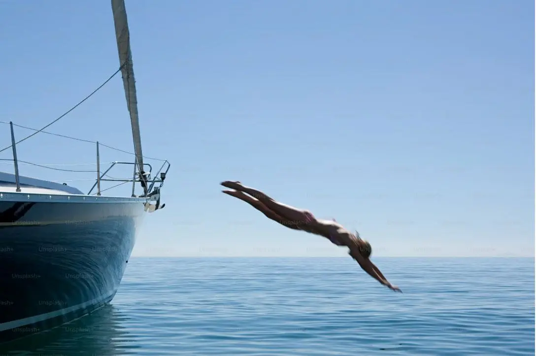 A woman in mid-dive gracefully plunges into the calm, clear sea from the bow of a sailing yacht on a sunny day.