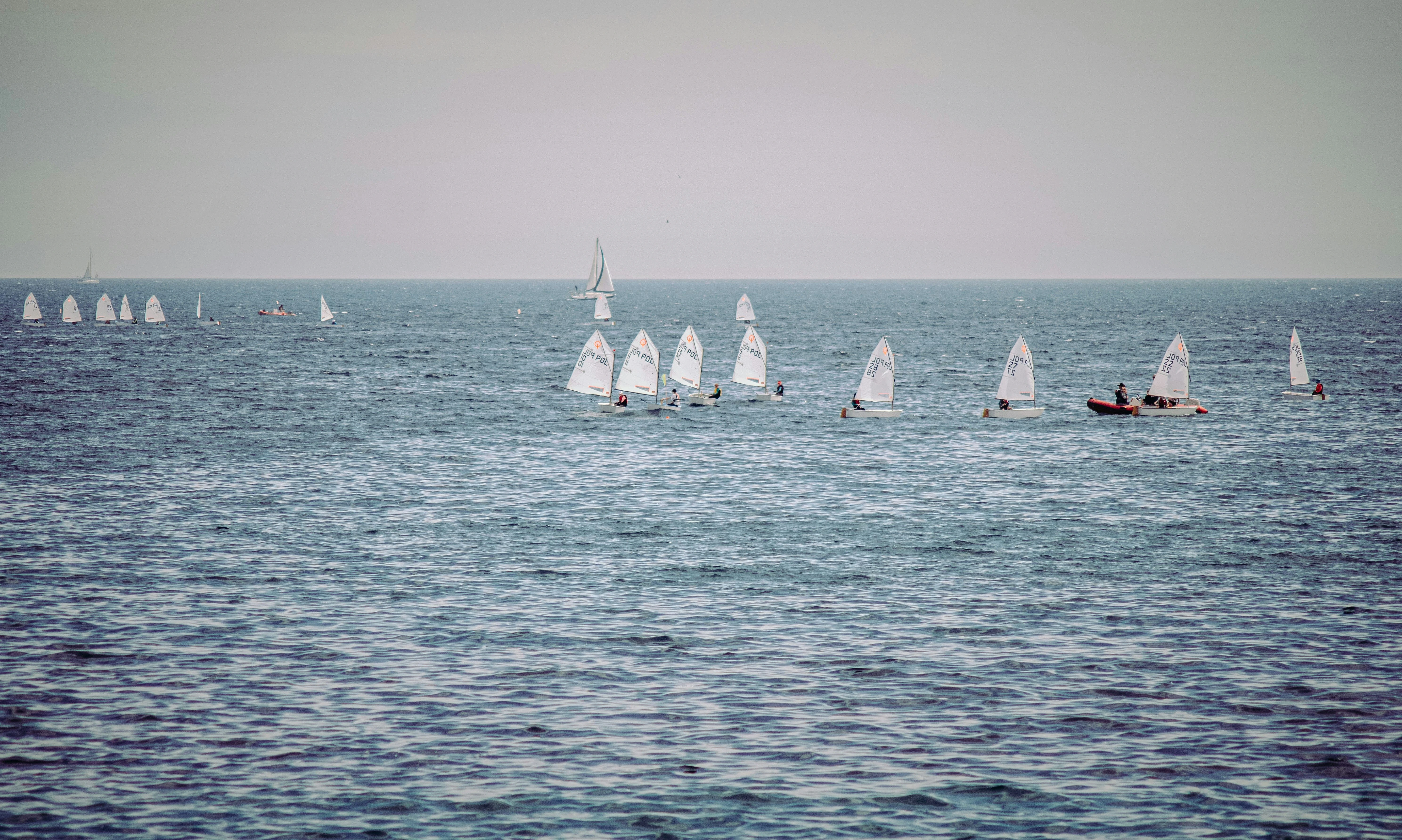 A group of small sailboats and a motorboat on the open sea, with some sailors sailing and others on the boat, under a clear sky.