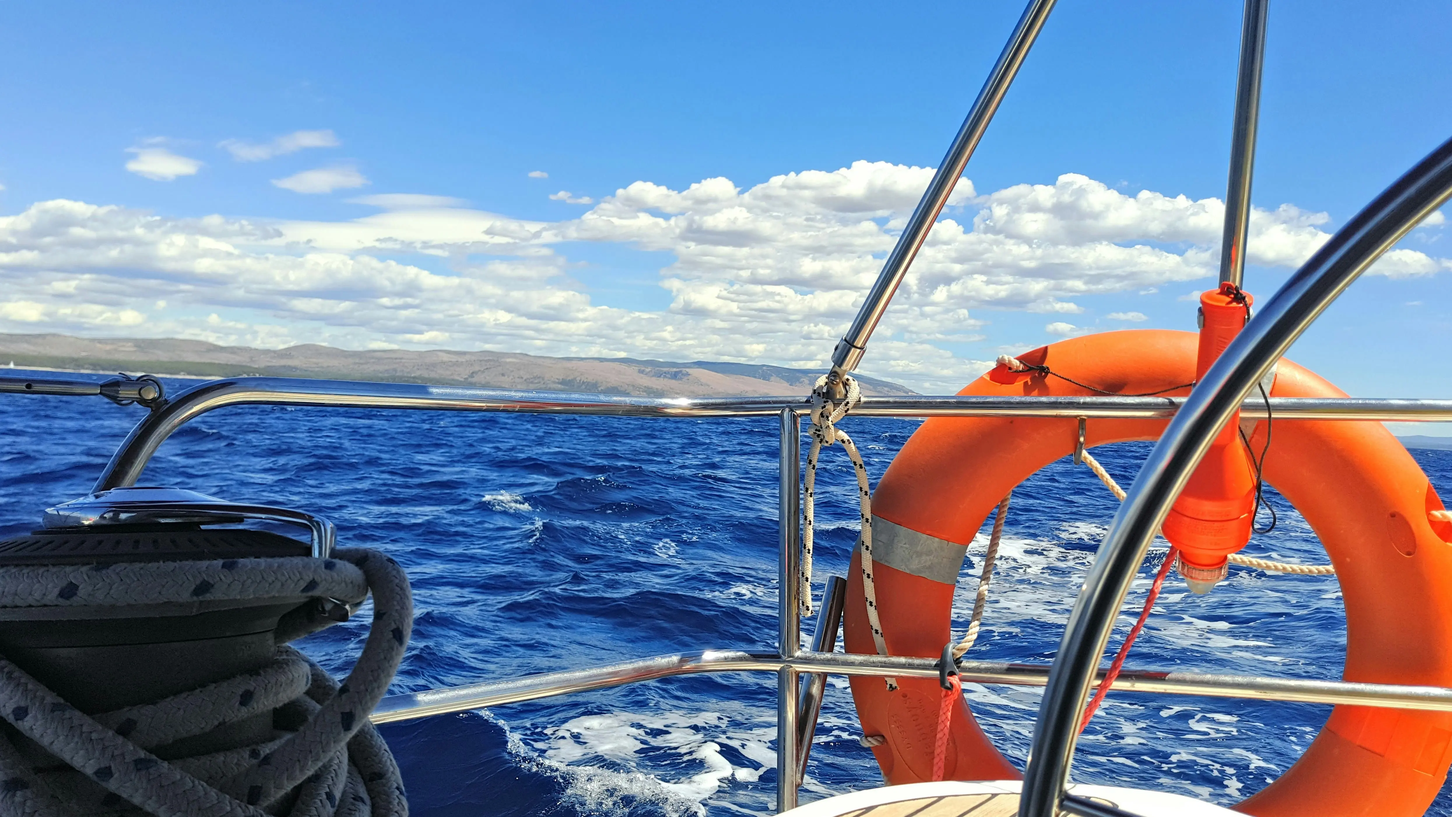 Aft view from a sailing yacht with a bright orange life buoy, coiled ropes, and foaming blue waves under a vivid sky with scattered clouds.
