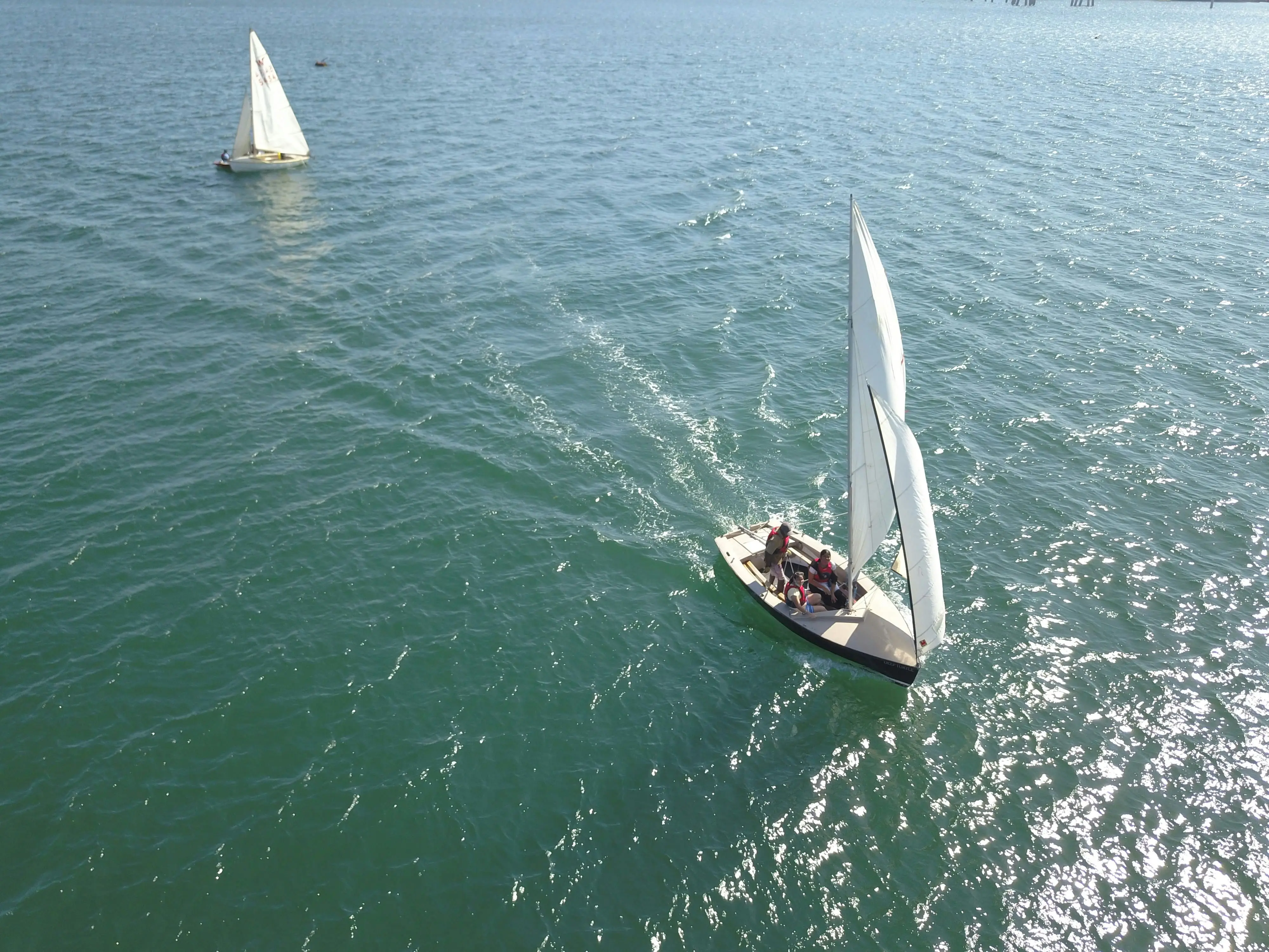 Aerial view of two small sailboats with people on board sailing on calm open water under a clear sky.