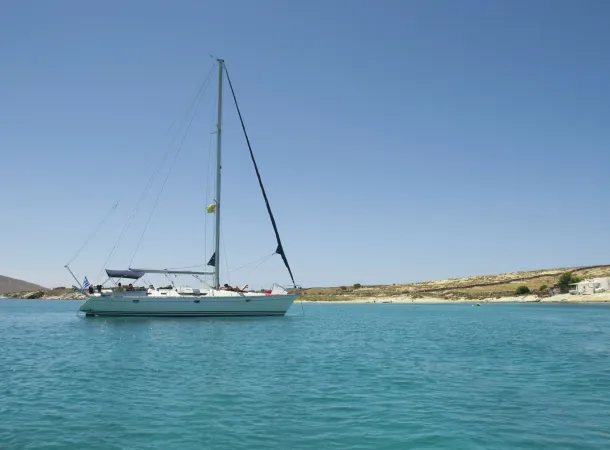 Sailing yacht anchored in a tranquil turquoise bay with a clear blue sky and a rocky Cycladic coastline in the background.