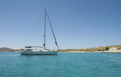Sailing yacht anchored in a tranquil turquoise bay with a clear blue sky and a rocky Cycladic coastline in the background.