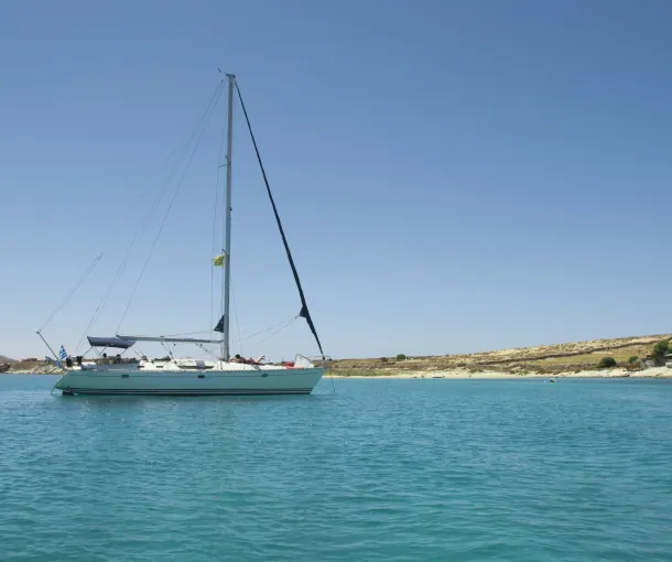 Sailing yacht anchored in a tranquil turquoise bay with a clear blue sky and a rocky Cycladic coastline in the background.