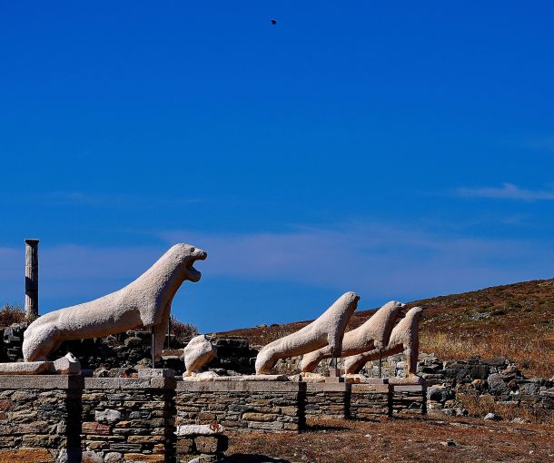 Ancient marble lion statues from the Terrace of the Lions stand in a row under a bright blue sky on Delos Island, Greece. Surrounded by weathered ruins and dry, rocky terrain, these iconic sculptures highlight the island's rich archaeological and mythological heritage.