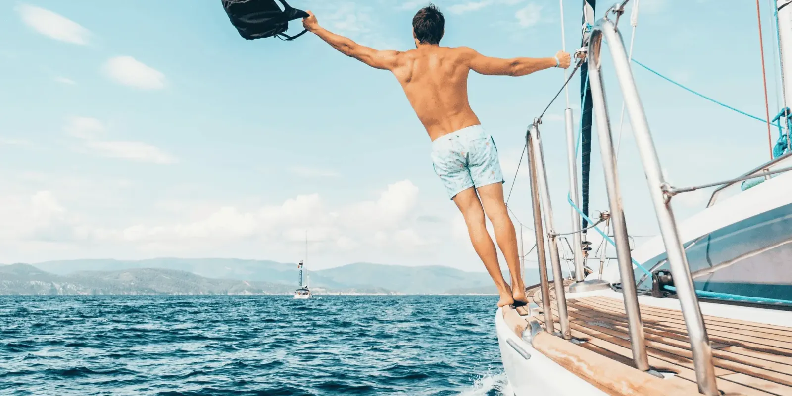 A man in swim trunks stands on the edge of a sailboat, holding a bag in one hand, with the ocean and distant shore in the background.