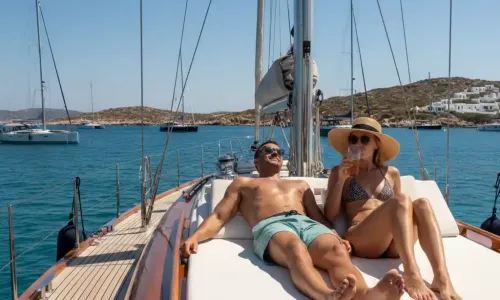 A couple relaxes on the deck of a sailing yacht anchored in a sunny Aegean Sea bay with other boats in the distance.