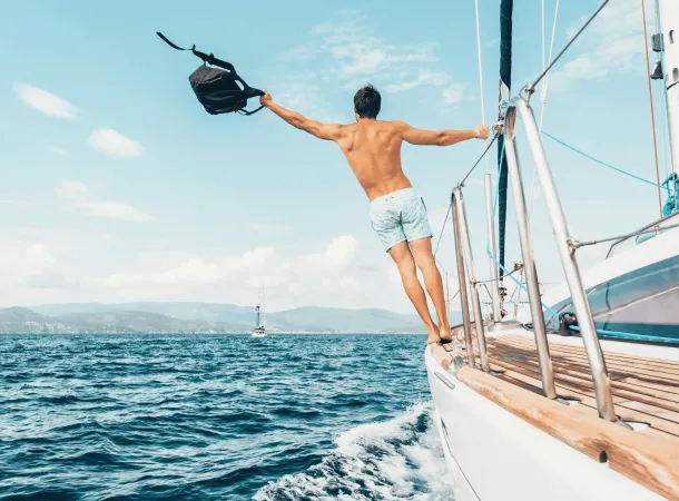 A man in swim trunks stands on the edge of a sailboat, holding a bag in one hand, with the ocean and distant shore in the background.