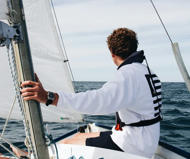 Young sailor wearing a white sweatshirt and life jacket, steering a small sailboat on open water, with sails hoisted and a calm horizon ahead.