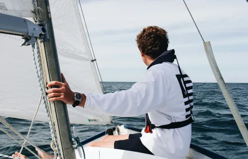 Young sailor wearing a white sweatshirt and life jacket, steering a small sailboat on open water, with sails hoisted and a calm horizon ahead.