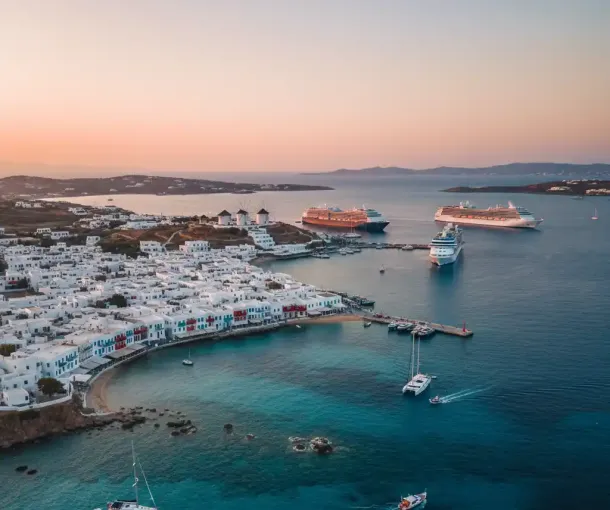 An aerial shot of Mykonos Town, showcasing its white buildings, coastal port, and several large cruise ships anchored in the bay during sunset.
