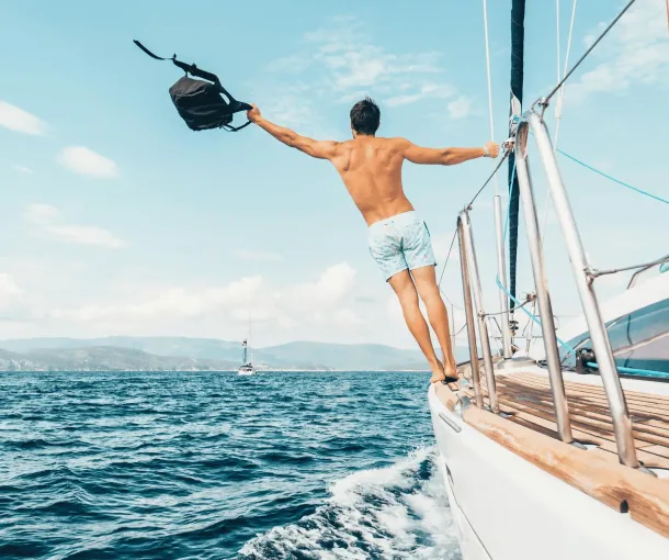 A man in swim trunks stands on the edge of a sailboat, holding a bag in one hand, with the ocean and distant shore in the background.