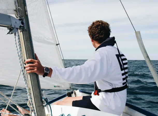 Young sailor wearing a white sweatshirt and life jacket, steering a small sailboat on open water, with sails hoisted and a calm horizon ahead.