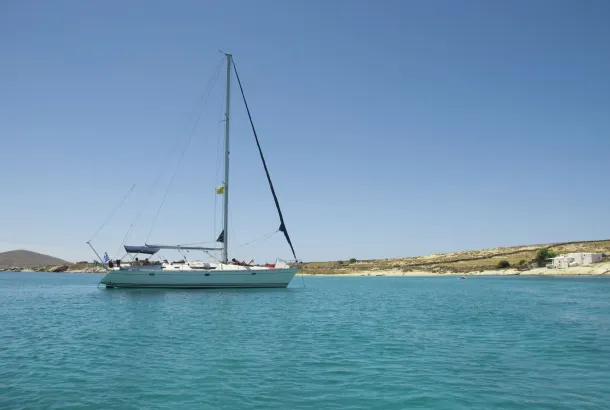 Sailing yacht anchored in a tranquil turquoise bay with a clear blue sky and a rocky Cycladic coastline in the background.