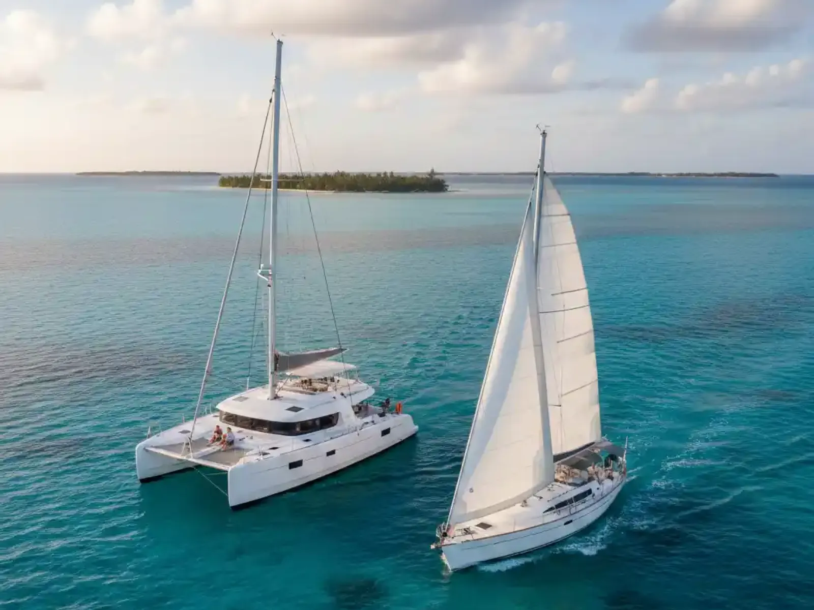 An aerial view of a white catamaran and a white monohull sailboat sailing side-by-side on clear turquoise waters, with a tropical island in the background under a partly cloudy sky at sunset.