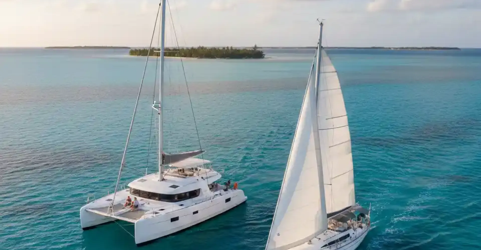 An aerial view of a white catamaran and a white monohull sailboat sailing side-by-side on clear turquoise waters, with a tropical island in the background under a partly cloudy sky at sunset.