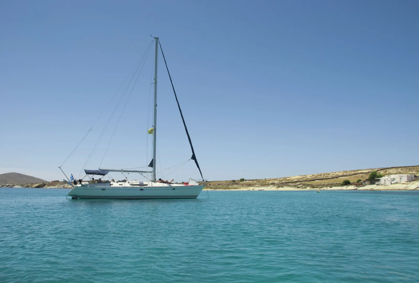 Sailing yacht anchored in a tranquil turquoise bay with a clear blue sky and a rocky Cycladic coastline in the background.