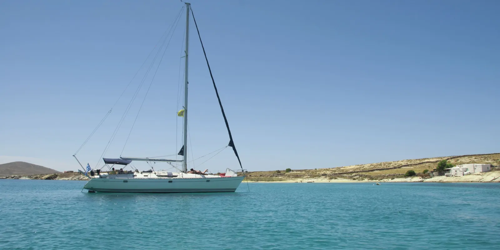 Sailing yacht anchored in a tranquil turquoise bay with a clear blue sky and a rocky Cycladic coastline in the background.