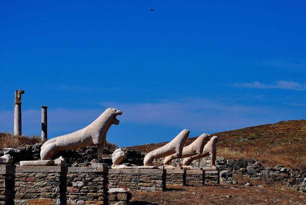 Ancient marble lion statues from the Terrace of the Lions stand in a row under a bright blue sky on Delos Island, Greece. Surrounded by weathered ruins and dry, rocky terrain, these iconic sculptures highlight the island's rich archaeological and mythological heritage.