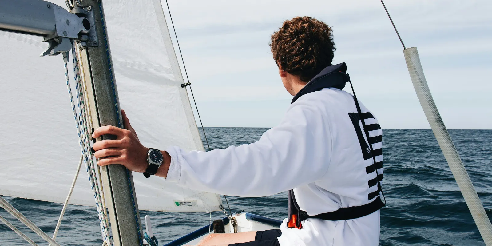 Young sailor wearing a white sweatshirt and life jacket, steering a small sailboat on open water, with sails hoisted and a calm horizon ahead.