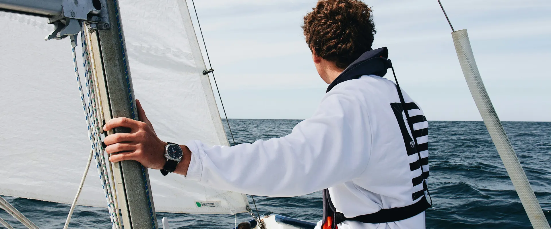 Young sailor wearing a white sweatshirt and life jacket, steering a small sailboat on open water, with sails hoisted and a calm horizon ahead.