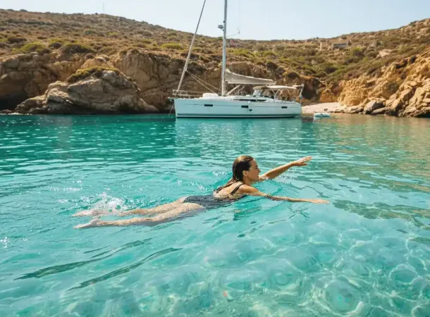 A person swimming in crystal clear turquoise waters off a beautiful, secluded cove, with a yacht visible in the background.