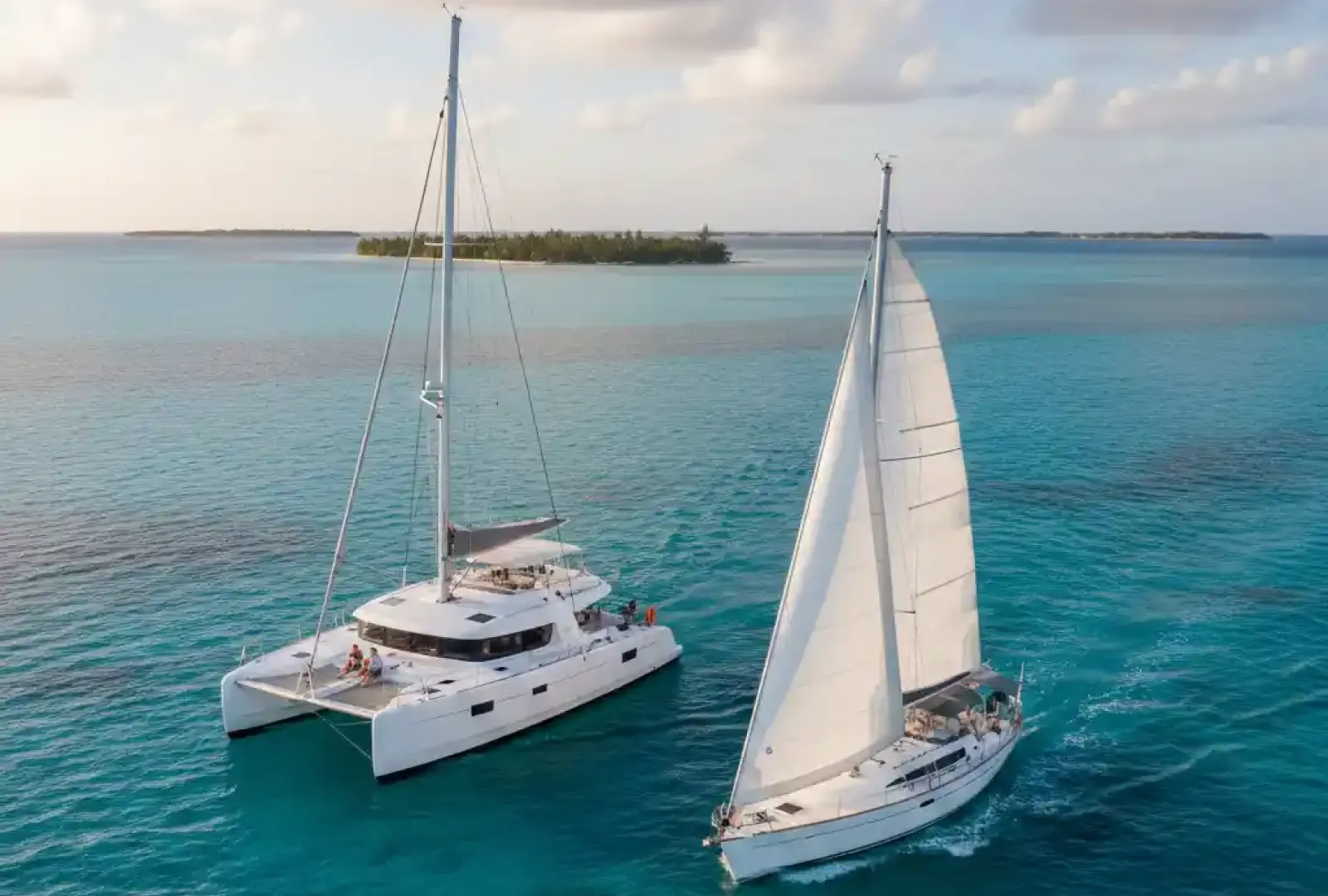An aerial view of a white catamaran and a white monohull sailboat sailing side-by-side on clear turquoise waters, with a tropical island in the background under a partly cloudy sky at sunset.