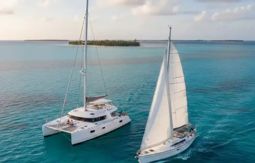 An aerial view of a white catamaran and a white monohull sailboat sailing side-by-side on clear turquoise waters, with a tropical island in the background under a partly cloudy sky at sunset.