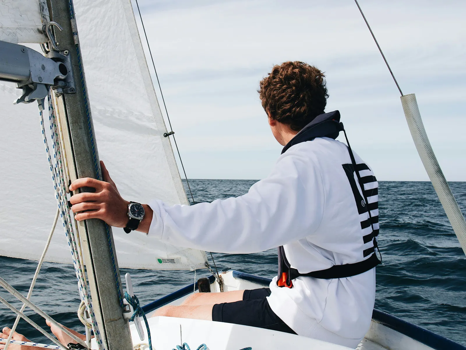 Young sailor wearing a white sweatshirt and life jacket, steering a small sailboat on open water, with sails hoisted and a calm horizon ahead.