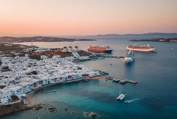 An aerial shot of Mykonos Town, showcasing its white buildings, coastal port, and several large cruise ships anchored in the bay during sunset.