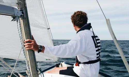 Young sailor wearing a white sweatshirt and life jacket, steering a small sailboat on open water, with sails hoisted and a calm horizon ahead.