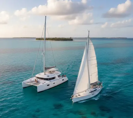 An aerial view of a white catamaran and a white monohull sailboat sailing side-by-side on clear turquoise waters, with a tropical island in the background under a partly cloudy sky at sunset.