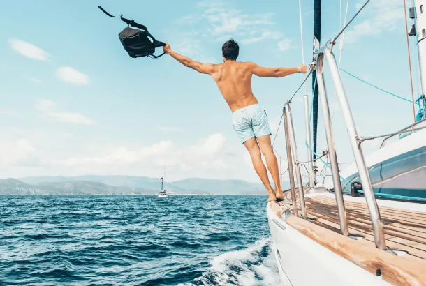 A man in swim trunks stands on the edge of a sailboat, holding a bag in one hand, with the ocean and distant shore in the background.