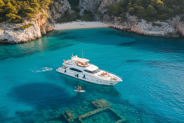 An aerial view of a luxury yacht anchored in a secluded, turquoise-water bay with pine-covered cliffs and a small beach, with a person paddleboarding near underwater ruins.