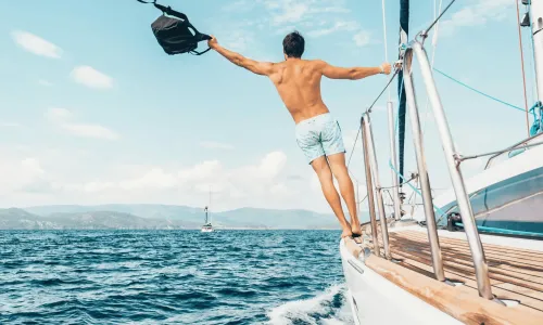 A man in swim trunks stands on the edge of a sailboat, holding a bag in one hand, with the ocean and distant shore in the background.