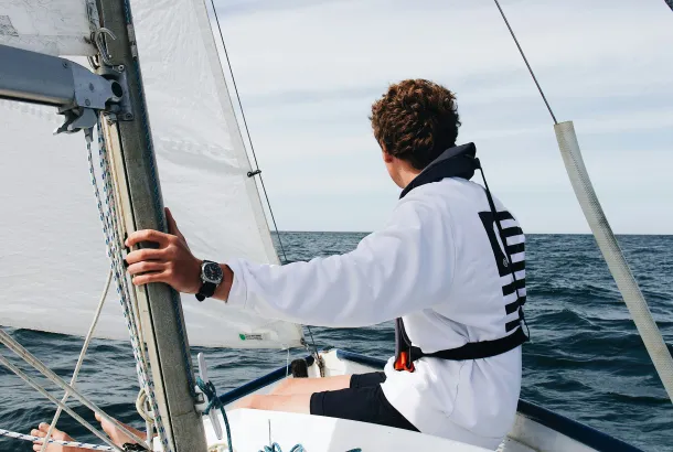 Young sailor wearing a white sweatshirt and life jacket, steering a small sailboat on open water, with sails hoisted and a calm horizon ahead.