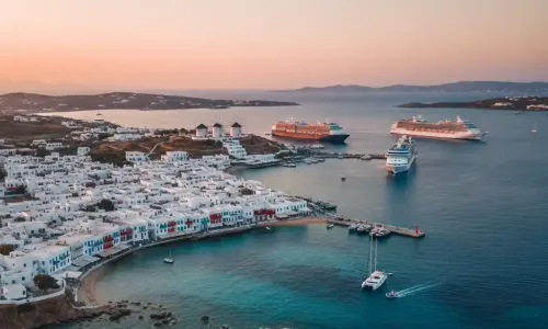 An aerial shot of Mykonos Town, showcasing its white buildings, coastal port, and several large cruise ships anchored in the bay during sunset.