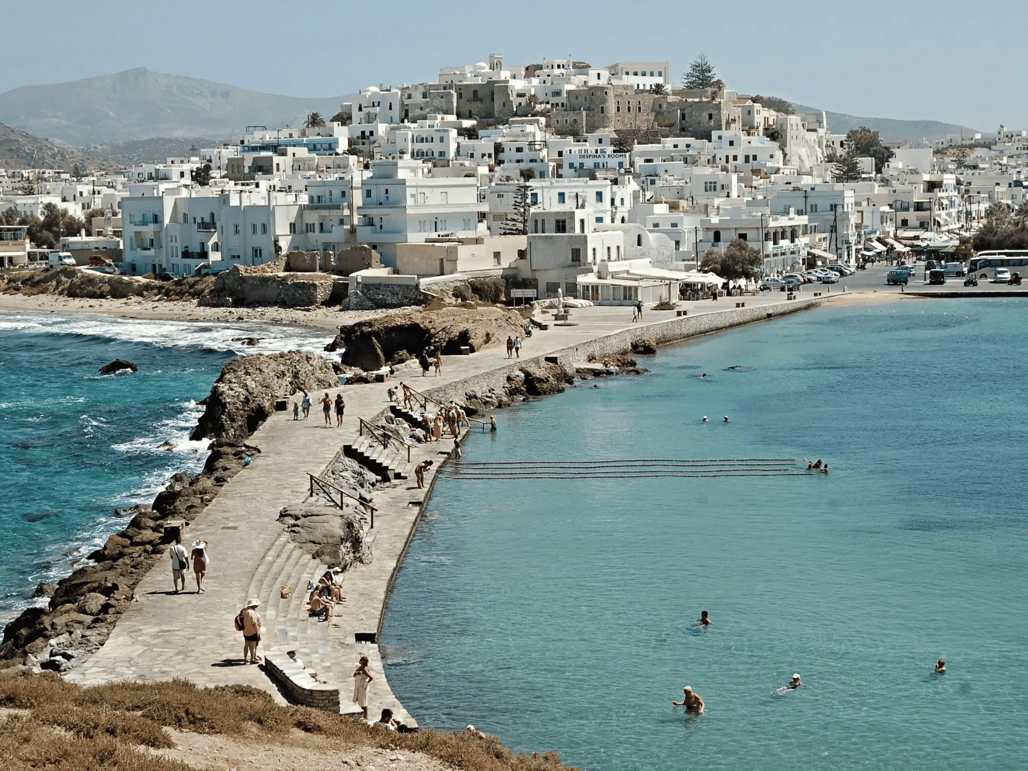 Seaside view of a whitewashed Greek town with a stone walkway, swimmers in turquoise water, and hills in the background.