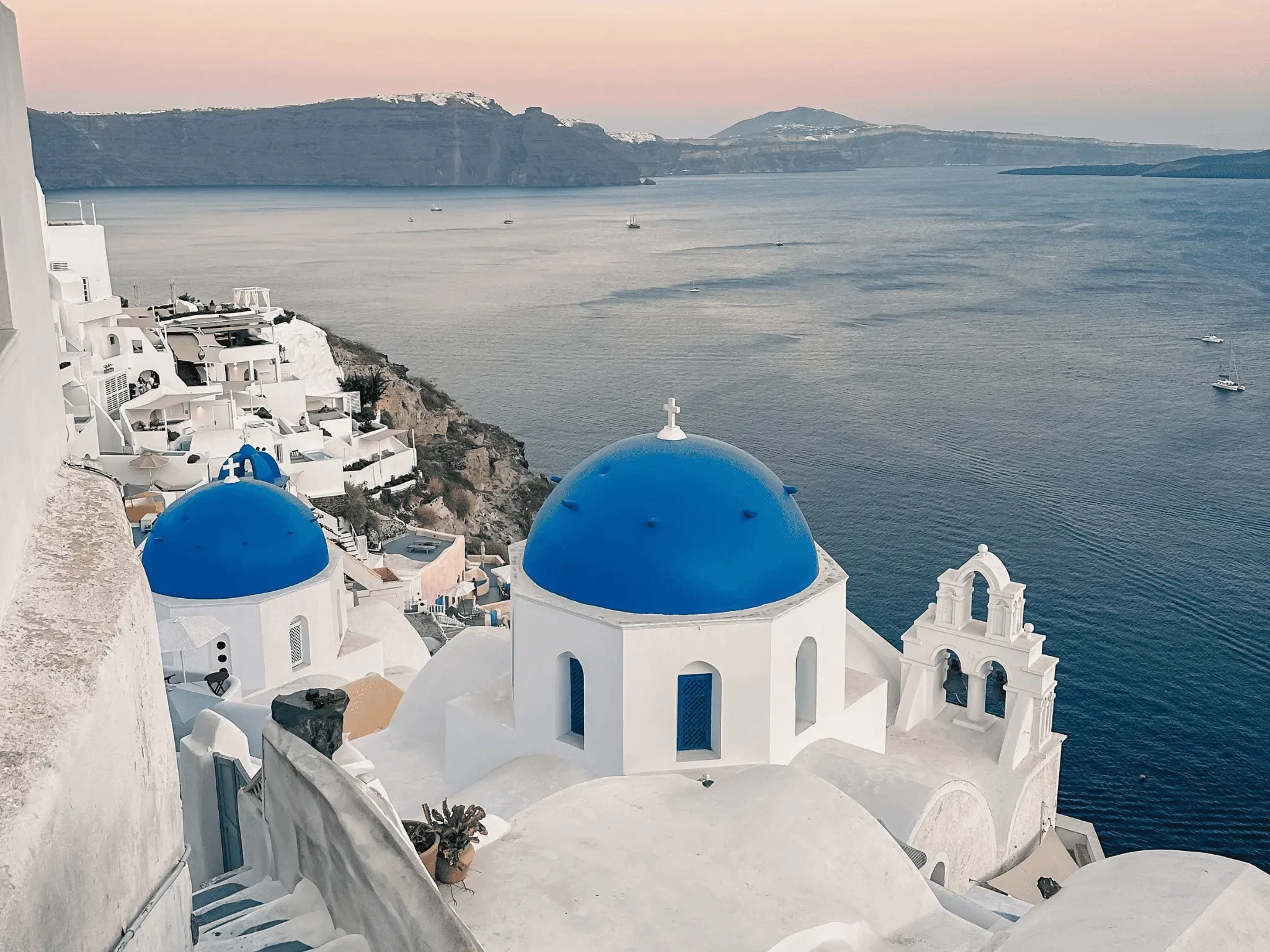 Blue-domed white churches in Oia, Santorini overlooking the caldera and Aegean Sea at sunset.