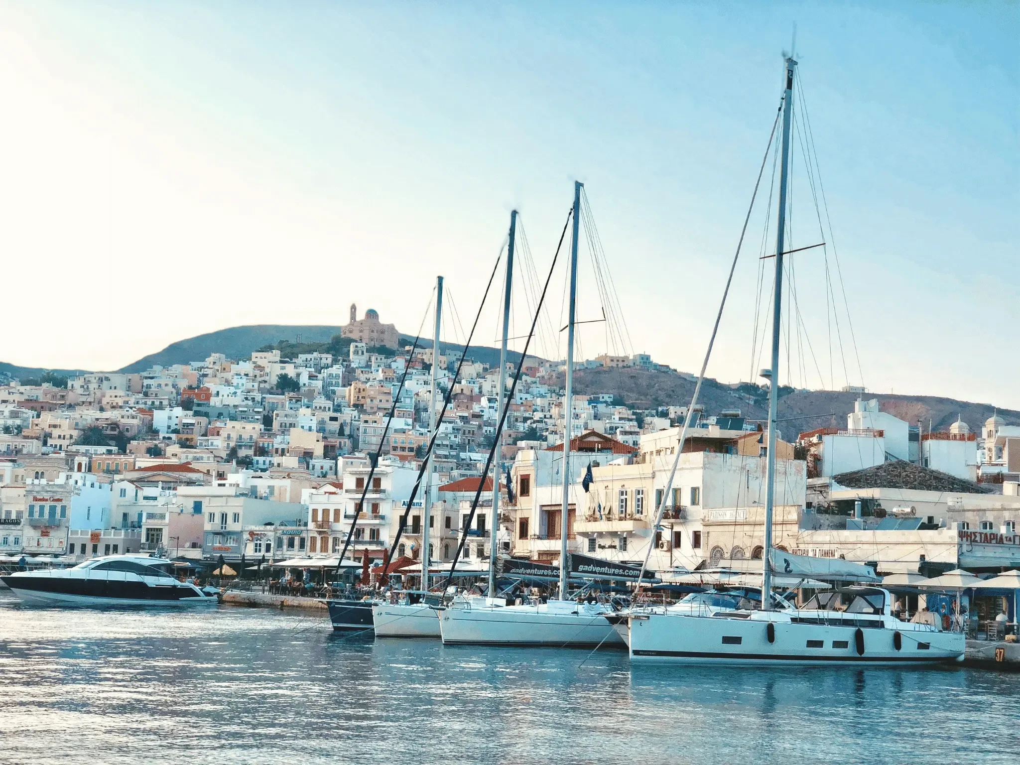Sailing yachts moored in Ermoupoli harbor, Syros Island, Greece with neoclassical hillside town in the background.