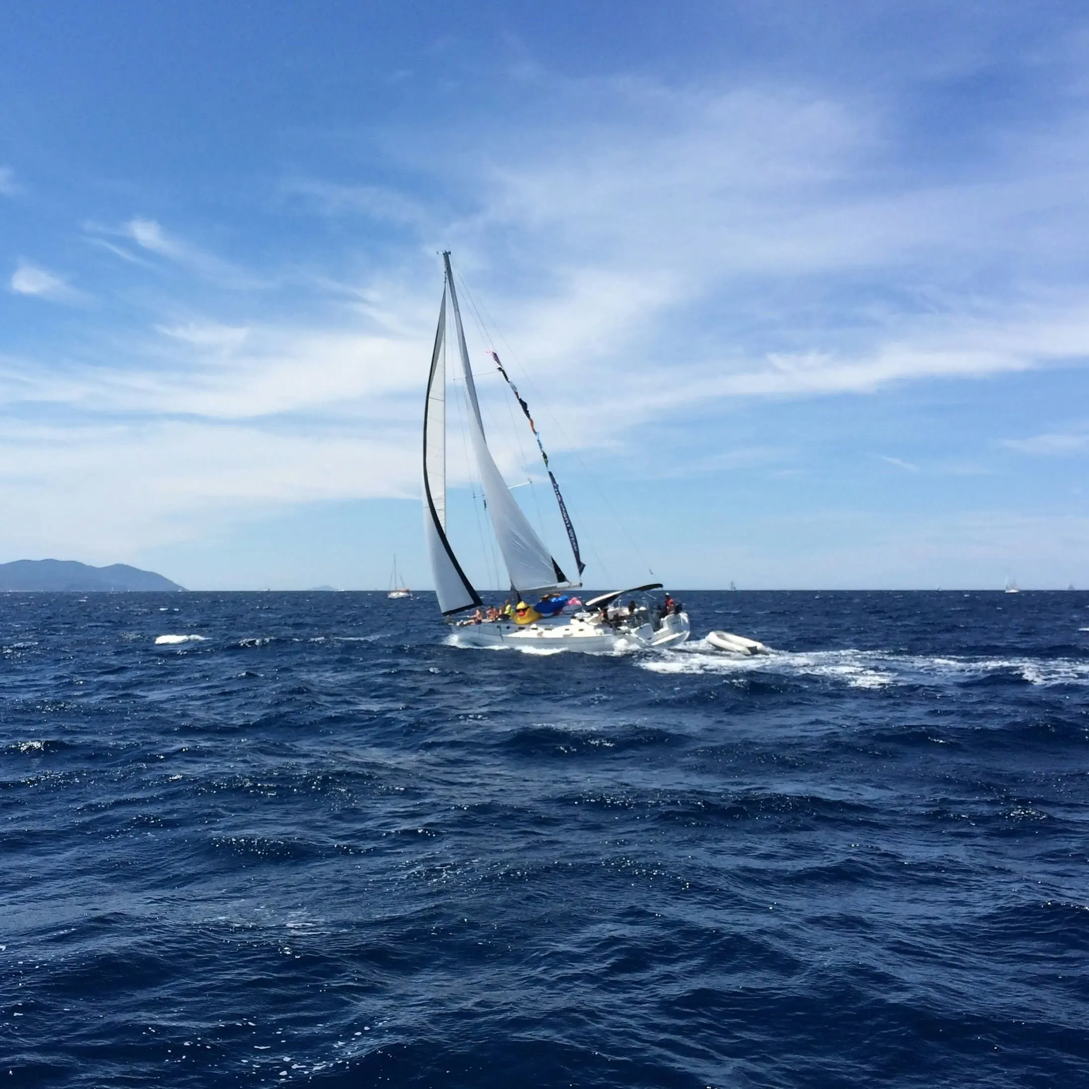 A sailing yacht with full sails glides through deep blue waters on a clear, sunny day with scattered clouds and distant mountains on the horizon.