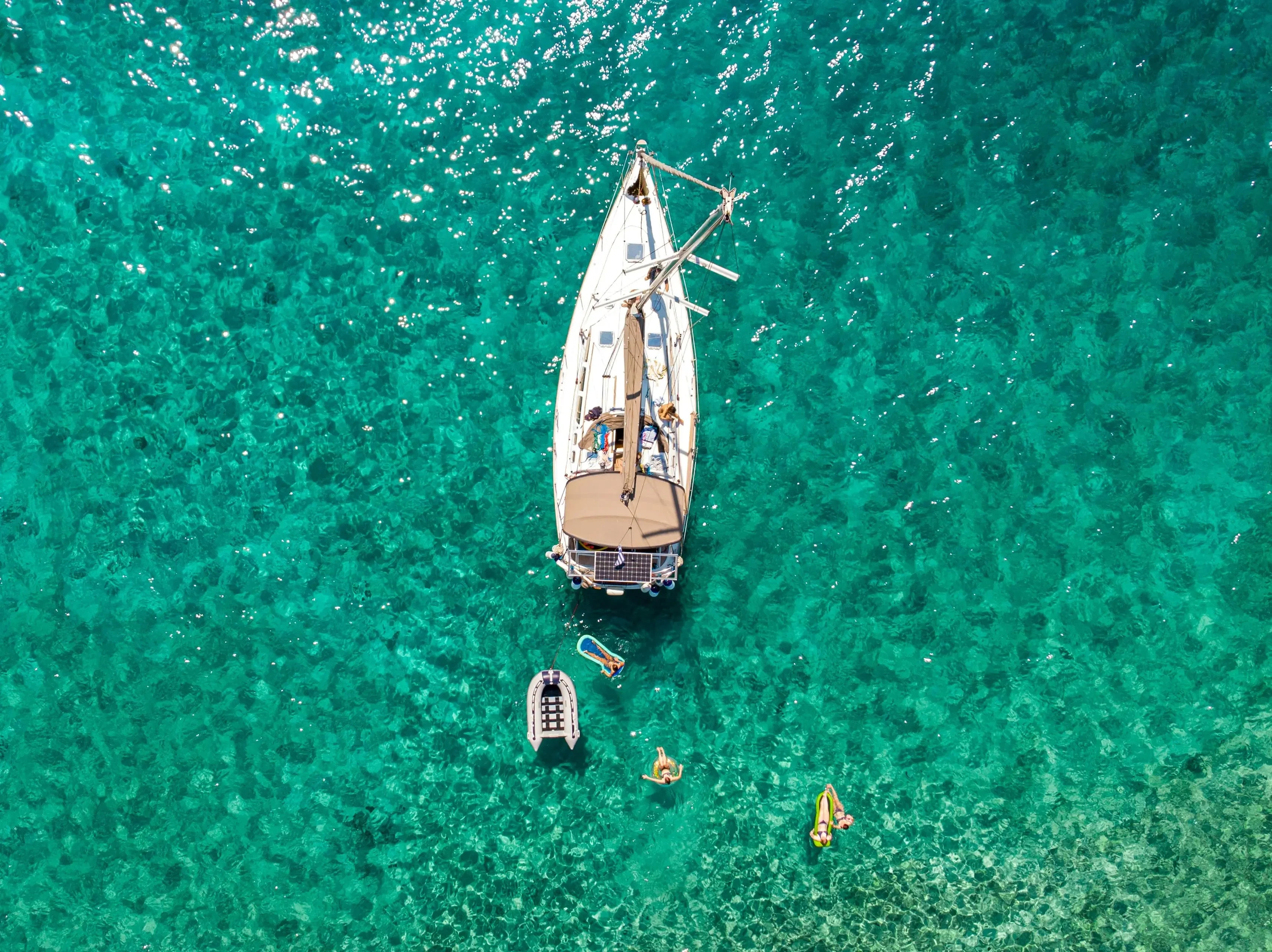 Aerial view of a sailing yacht anchored in crystal-clear turquoise waters with people swimming and relaxing on floaties nearby.